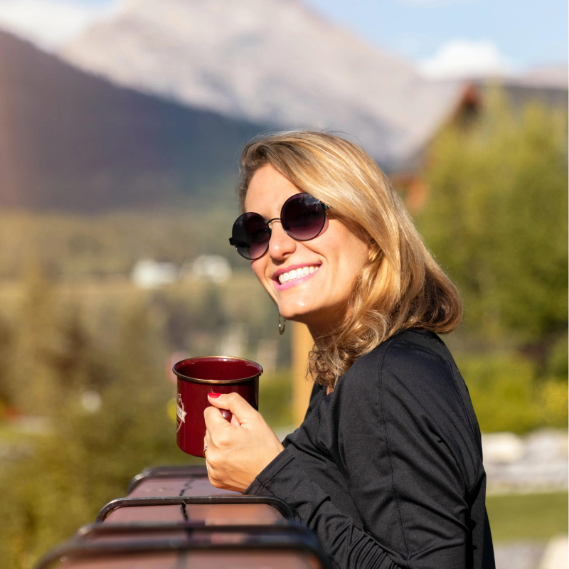 Smiling woman with sunglasses and coffee cup standing on balcony in mountains